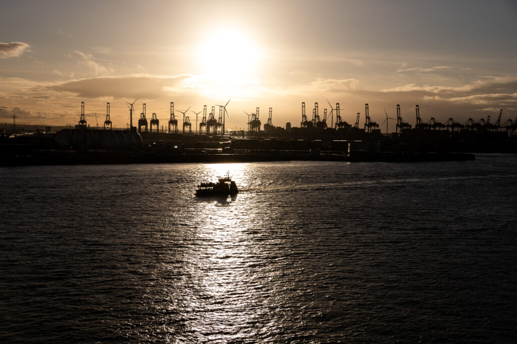 Ein goldener Sonnenuntergang über dem Hamburg Hafen, während die Silhouette eines kleinen Bootes auf dem Wasser zu sehen ist.